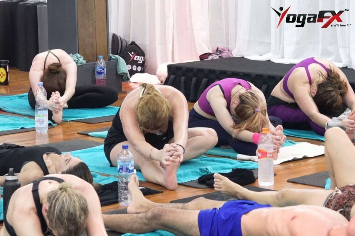 What to bring to hot yoga: students in a seated forward bend on teal towels with water bottles beside them, wearing fitted sports tops and shorts at a YogaFX class in Bali